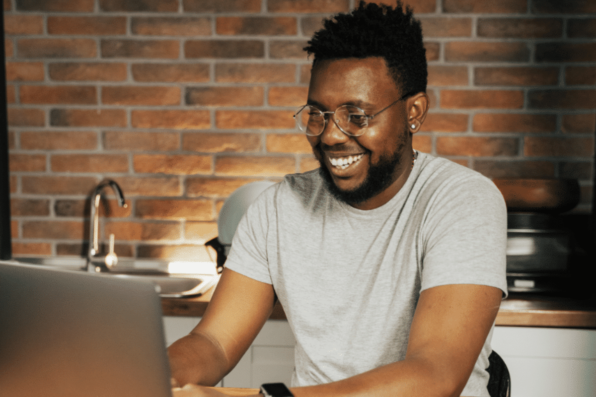 Jovem negro sorridente digitando no notebook enquanto faz curso online em casa na cozinha