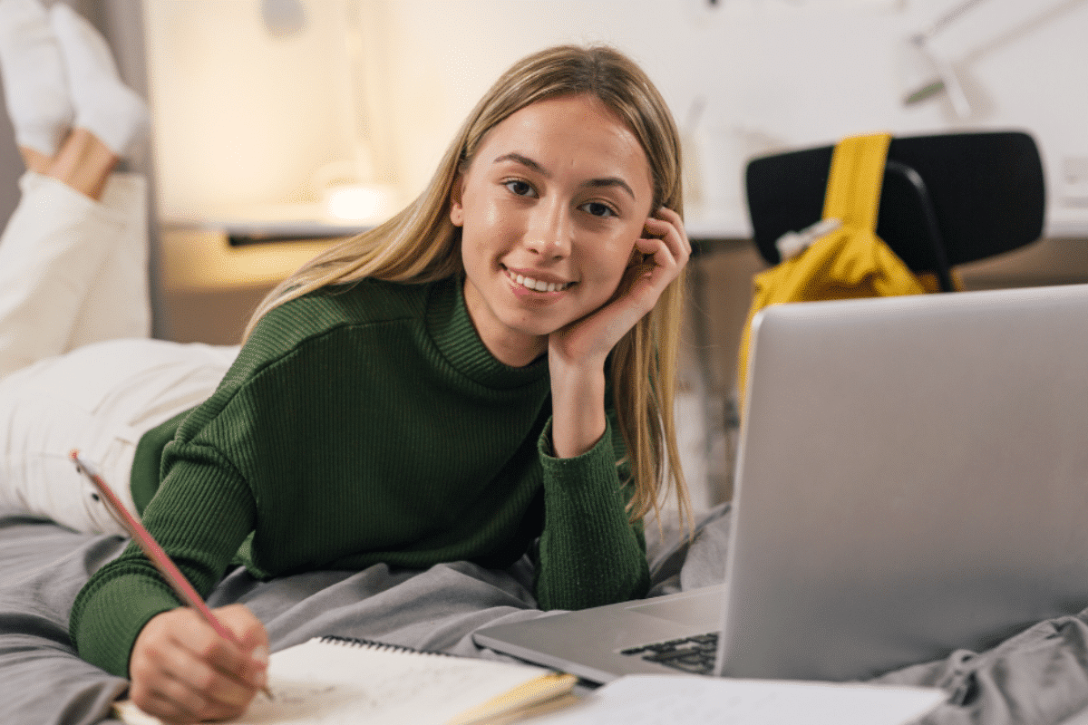 Jovem garota sorridente estudando cursos online, deitada na cama enquanto anota no caderno e usa notebook