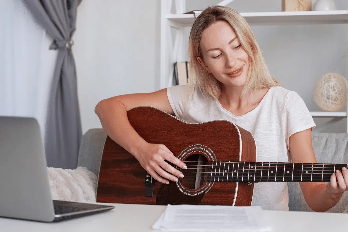 Jovem loira sorridente aprendendo a tocar violão com cursos online, usando notebook no sofá