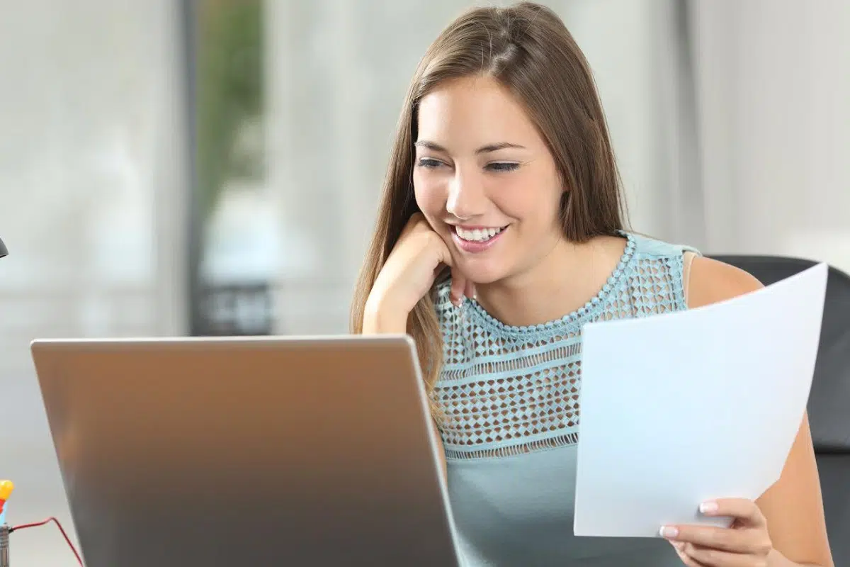 Mulher loira sorridente estudando cursos online, olhando para a tela do notebook, segurando uma folha de papel e apoiada na mesa