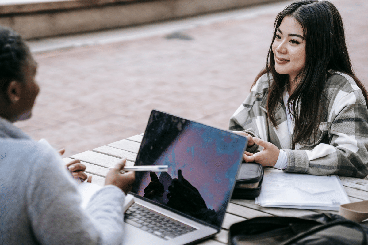 Duas jovens estudantes se preparando para entrevistas de emprego, estudando ao ar livre com notebook e materiais de estudo sobre a mesa