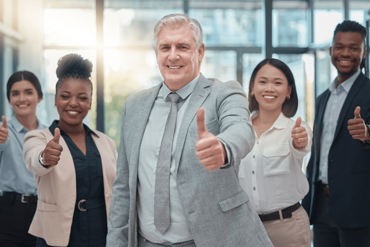 Cinco funcionários sorridentes em trajes sociais em ambiente corporativo, fazendo gesto de positivo, representando sucesso em entrevistas de emprego