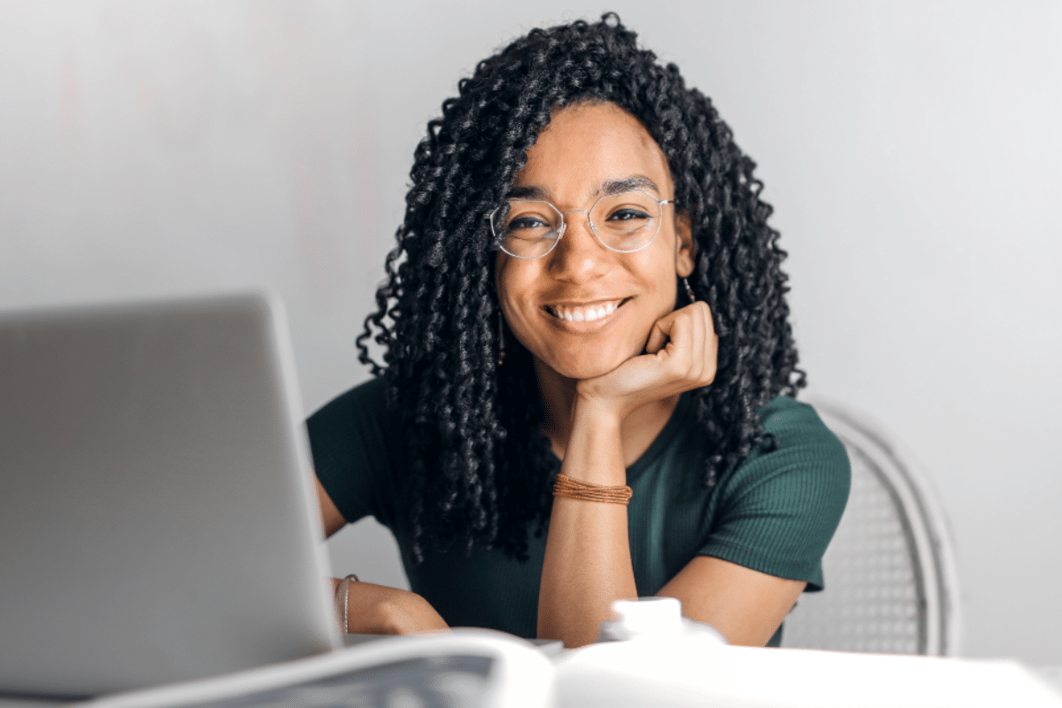 Jovem mulher morena sorridente de cabelo cacheado usando óculos de grau, estudando cursos que mais empregam no Brasil em casa, com notebook sobre a mesa