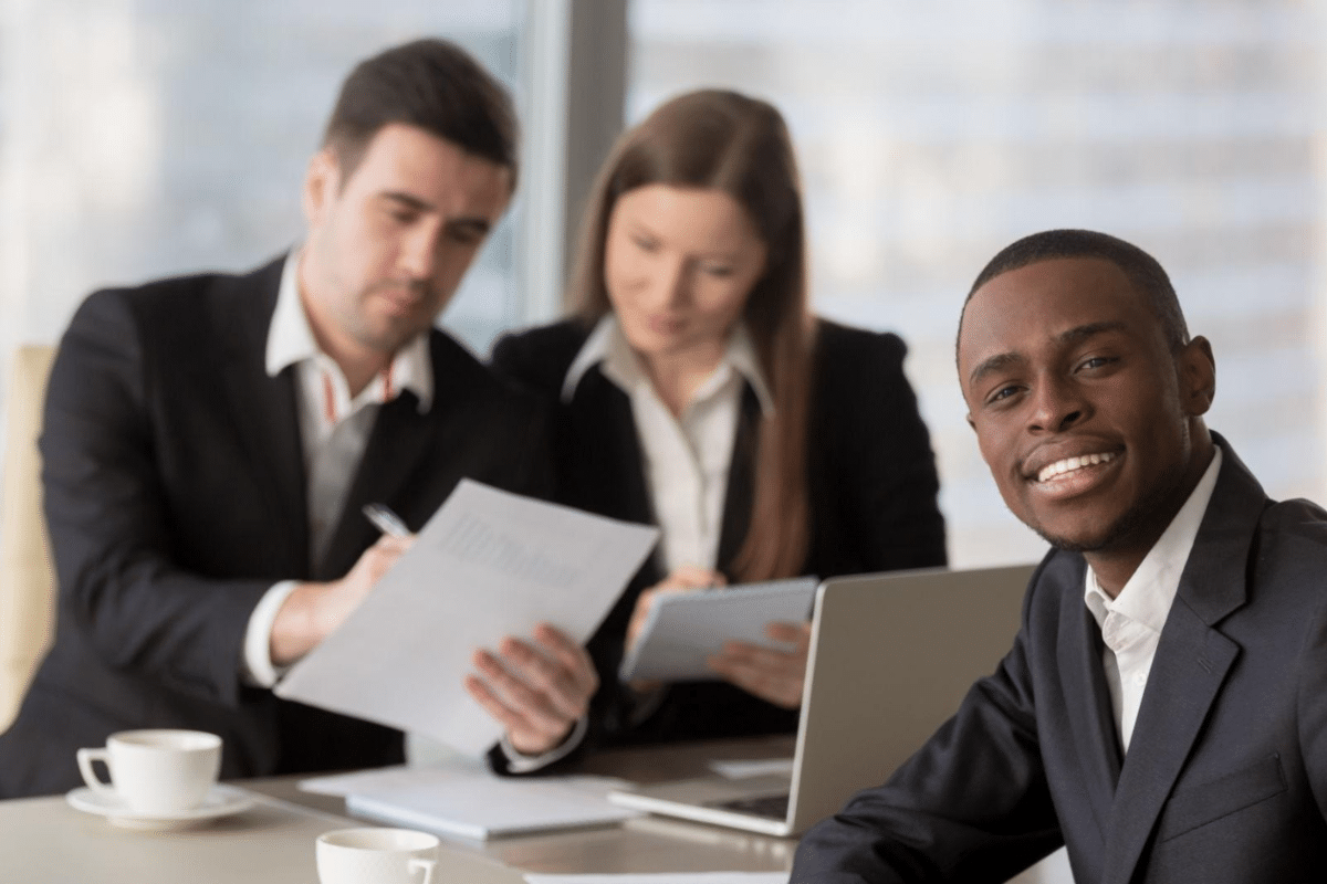 Jovem homem negro sorridente em entrevistas de emprego, sentado em cadeira, vestindo terno, sendo avaliado por dois empregadores em trajes sociais, com notebook e xícaras de café sobre a mesa em ambiente corporativo